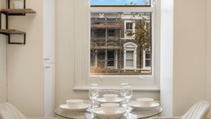 A dining area with a round glass table set for two, white chairs, and a window with a view of a building. - Cozy Home in Philbeach Garden (London)
