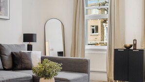 A modern living room with a grey sofa, a black coffee table, a large mirror, and a view of a building through a window. - Cozy Home in Philbeach Garden (London)