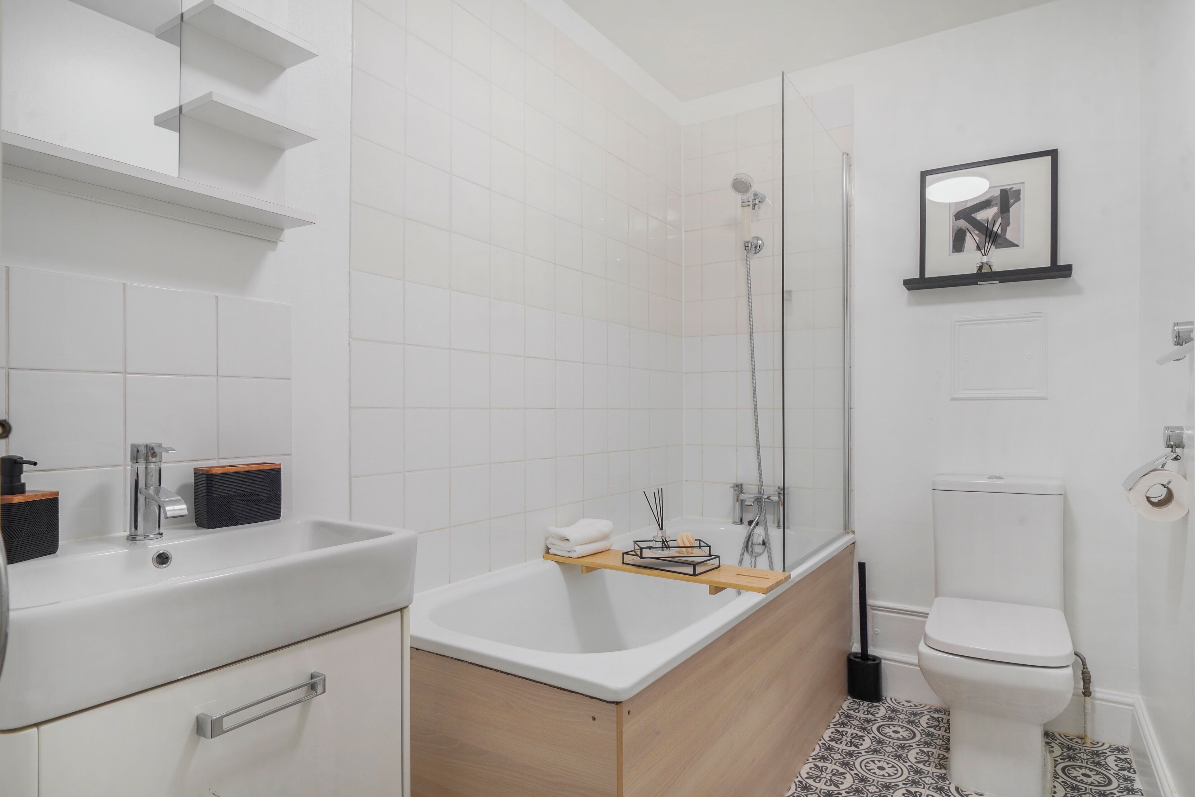 A modern bathroom with a white sink, a bathtub, and a toilet. There is a shelf with toiletries and a framed picture on the wall.