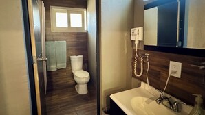 Un baño con suelo de madera, inodoro blanco, lavabo con espejo arriba y teléfono fijo en la pared.