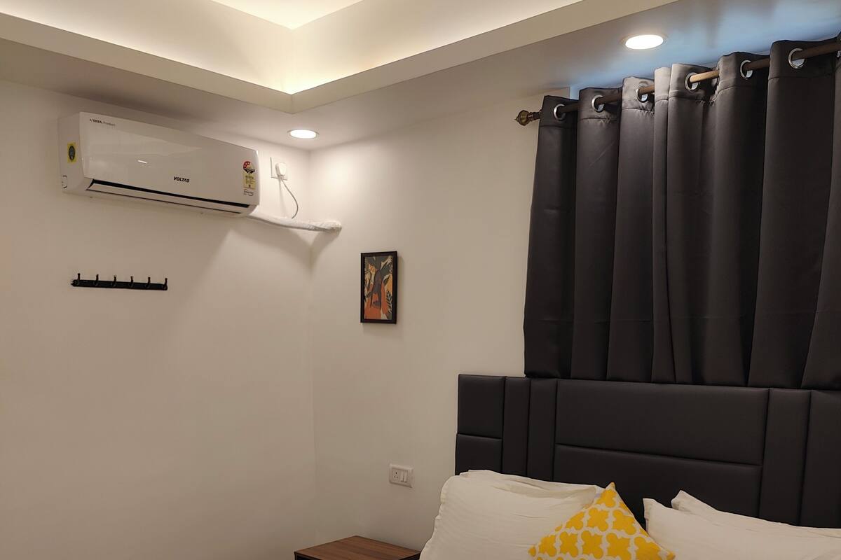 A hotel room with a large bed, a nightstand, a wall-mounted air conditioner, a small picture, and a window with dark curtains.