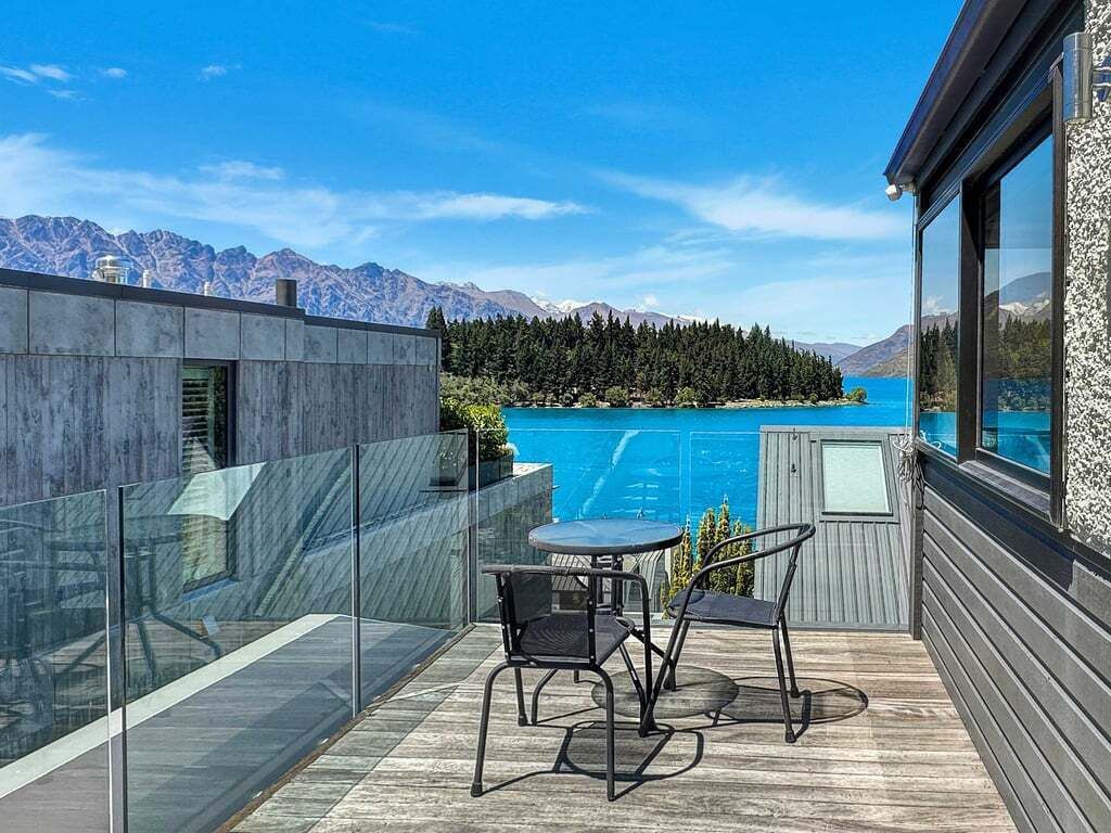 A balcony with a glass railing, a table, and two chairs, overlooking a lake and mountains.