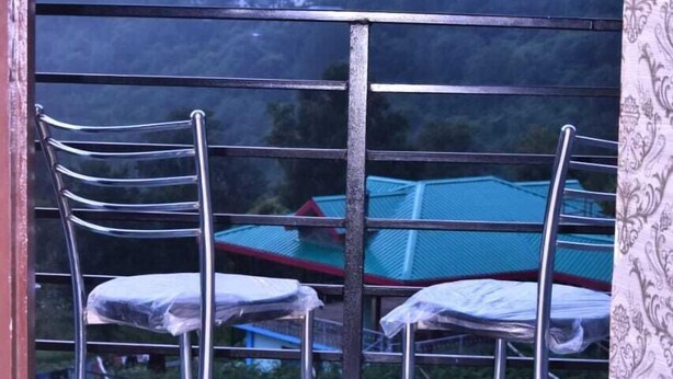 Ein Balkon mit Metallstühlen und einem Tisch, der auf eine grüne Landschaft mit Gebäuden im Hintergrund blickt.