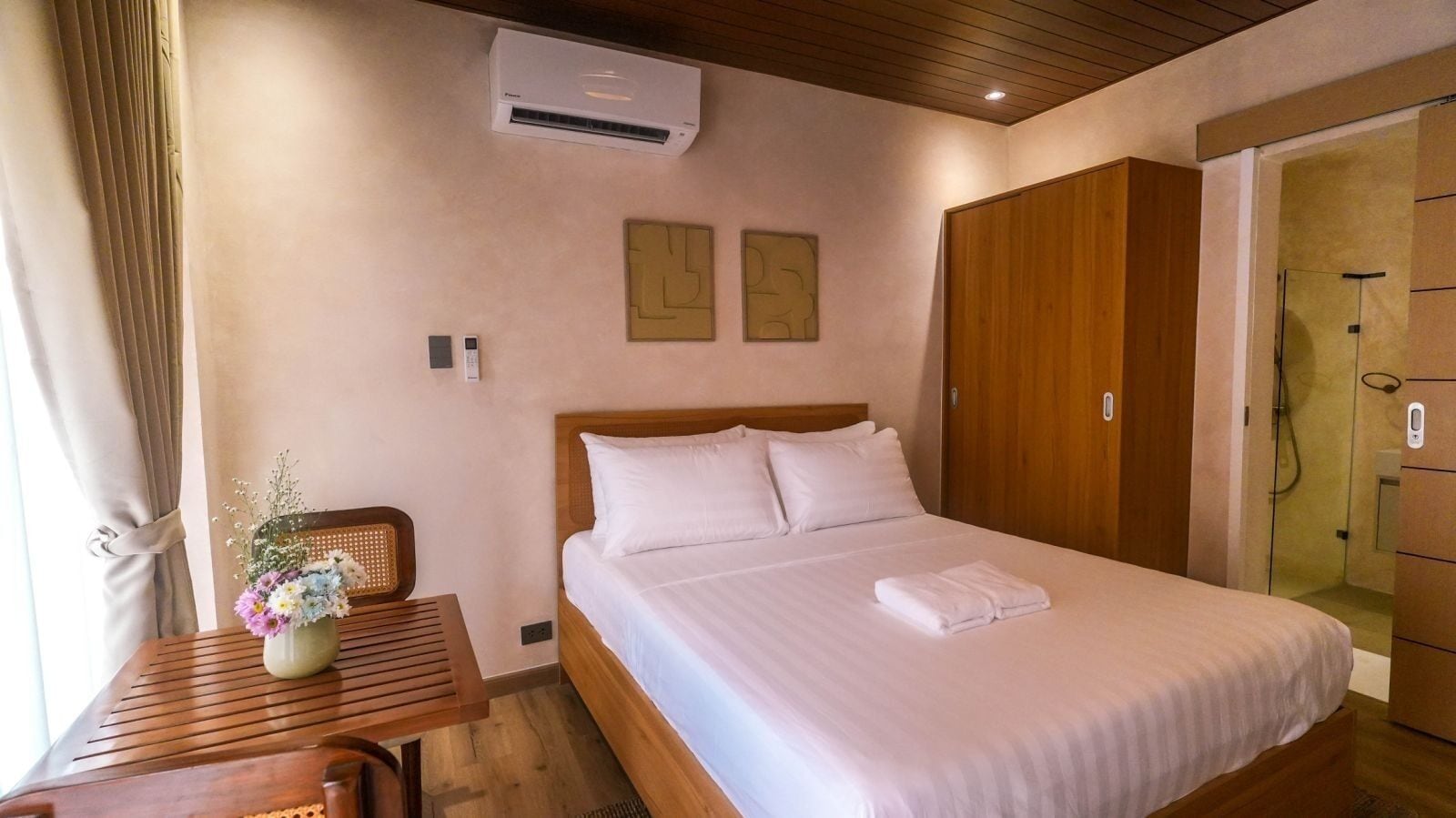 A neatly arranged hotel room with a bed, a wooden nightstand with a vase of flowers, a wall-mounted air conditioner, and a wooden wardrobe.