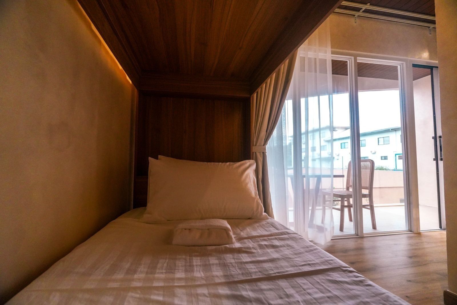 Una cama individual con cabecera de madera, una mesita de noche pequeña y vistas al exterior a través de un ventanal corredero.