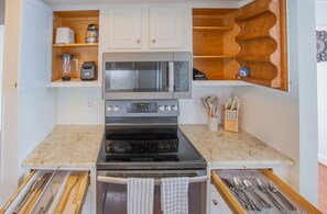 A compact kitchen with a stove, oven, microwave, and a built-in blender. There are wooden shelves above the stove and a knife block to the right. - HOA Townhouse in wonderful High Point by the Lake (High Point)