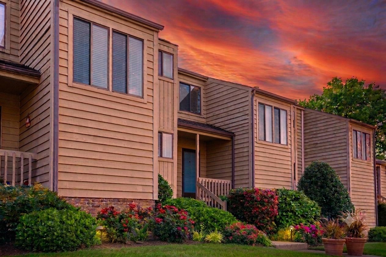 A row of two-story wooden townhouses with a front porch and a small garden area with various plants and flowers.