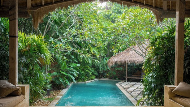 A pool surrounded by lush greenery, with a thatched roof structure and wooden seating areas. - Nomadic Resort Bali (Kerobokan)