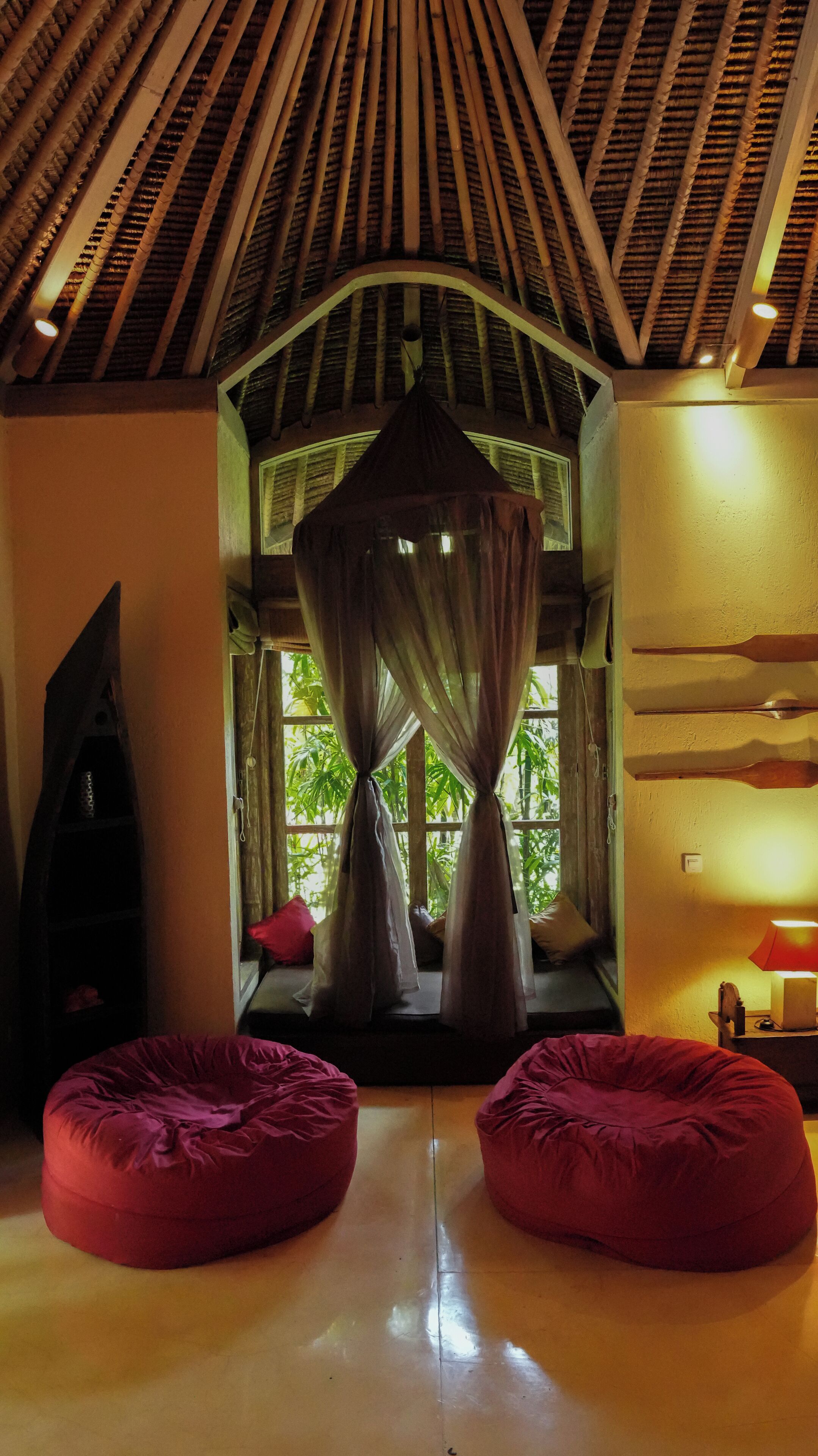 A room with a thatched roof, a bed with red cushions, and a large window with curtains.