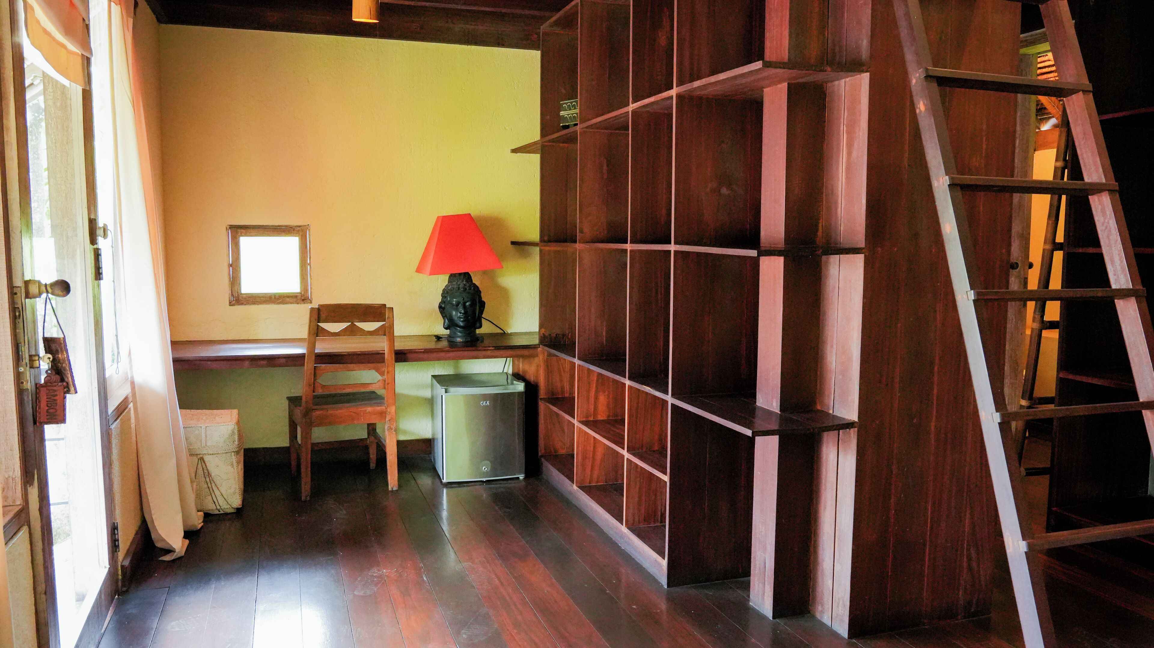 Una habitación con un escritorio de madera, una silla, una lámpara roja, una estantería y una escalera.