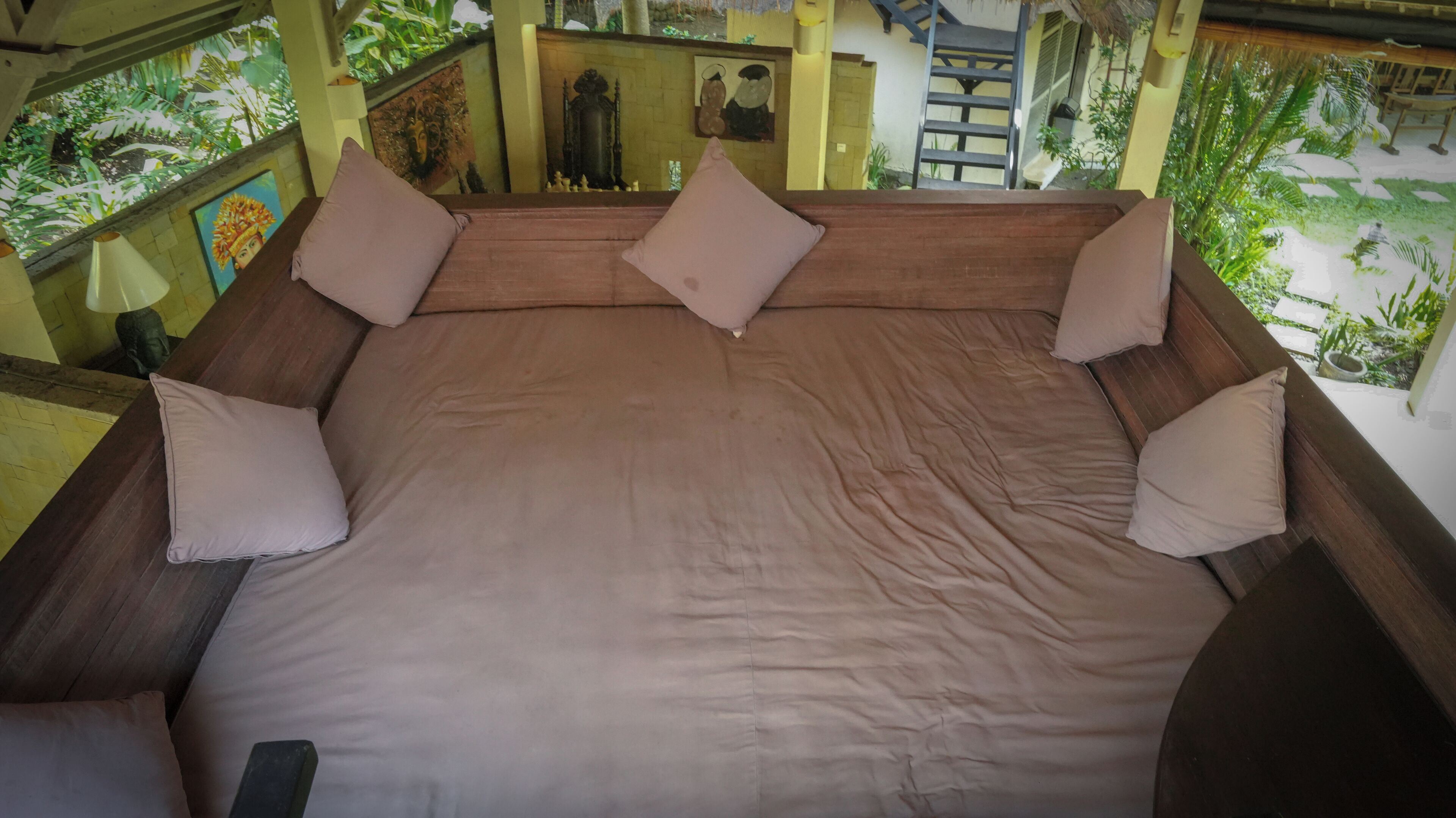 A large bed with a wooden frame and multiple pillows, set in a room with a view of greenery and a staircase.