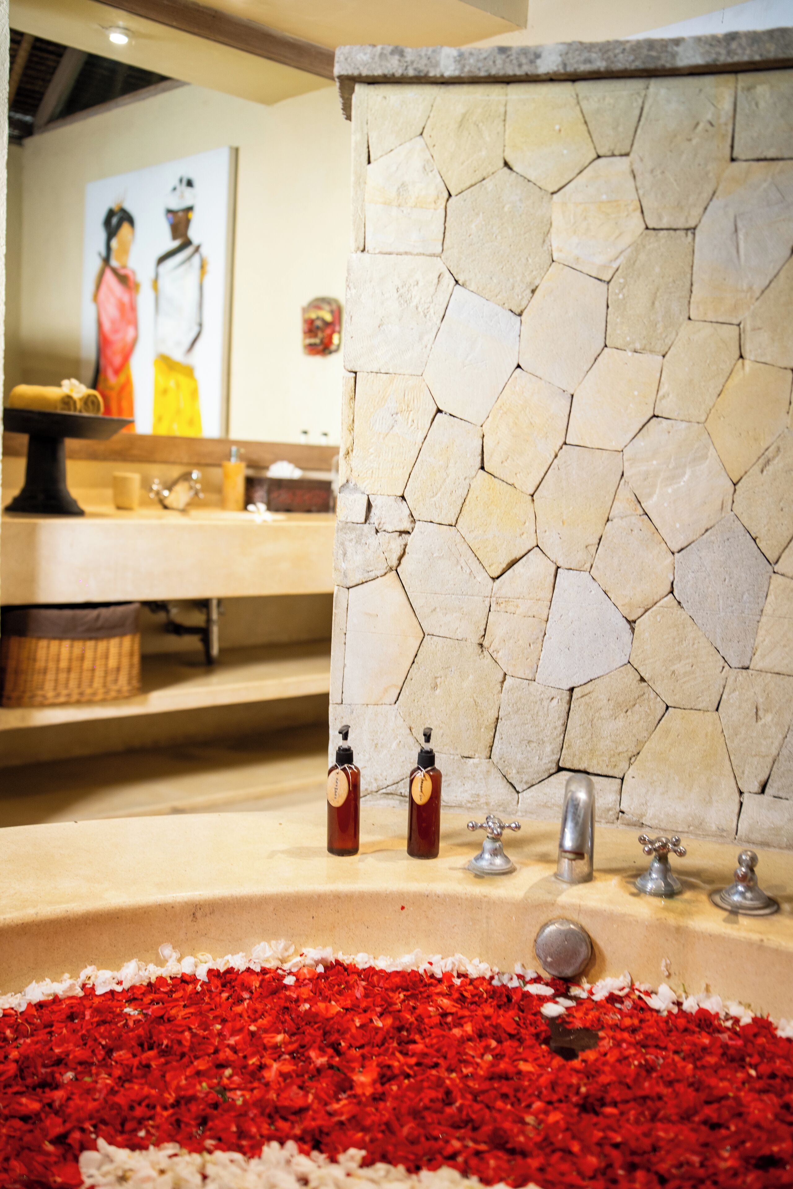 Un baño con pared de mosaicos de piedra, un lavamanos adornado con pétalos de rosa y un cuadro en la pared.
