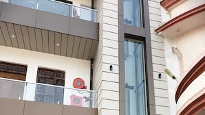 A multi-story building with a modern design, featuring a central glass elevator shaft, a fire alarm system, and a visible entrance with steps leading up to the door. - Hotel Pinaka (Pratapgarh)