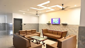 A modern lounge area with leather sofas, a glass coffee table, recessed ceiling lighting, a ceiling fan, and a flat-screen TV mounted on the wall. - Hotel Pinaka (Pratapgarh)