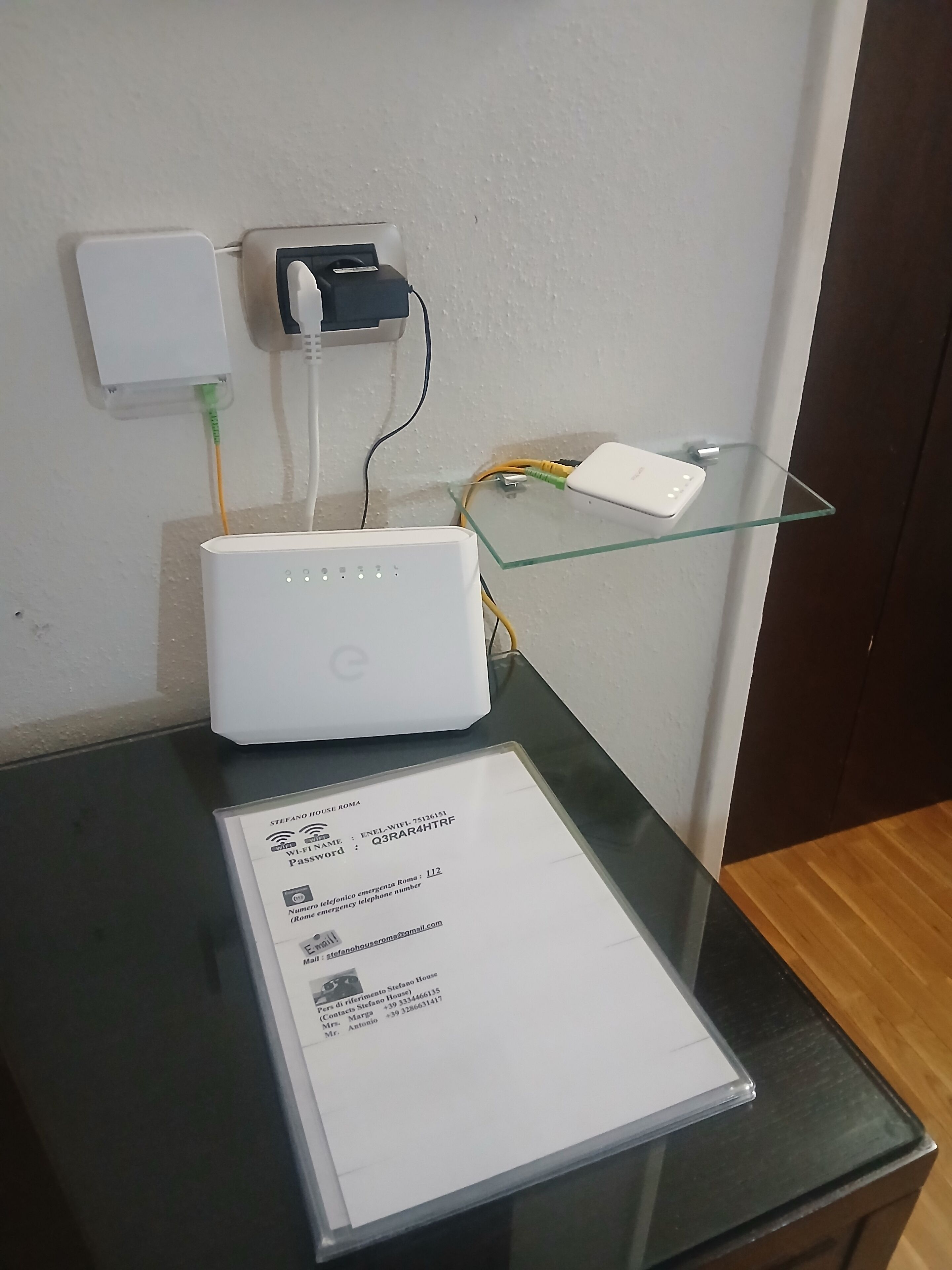 A white router on a glass shelf, a white power strip, and a document on a black table.