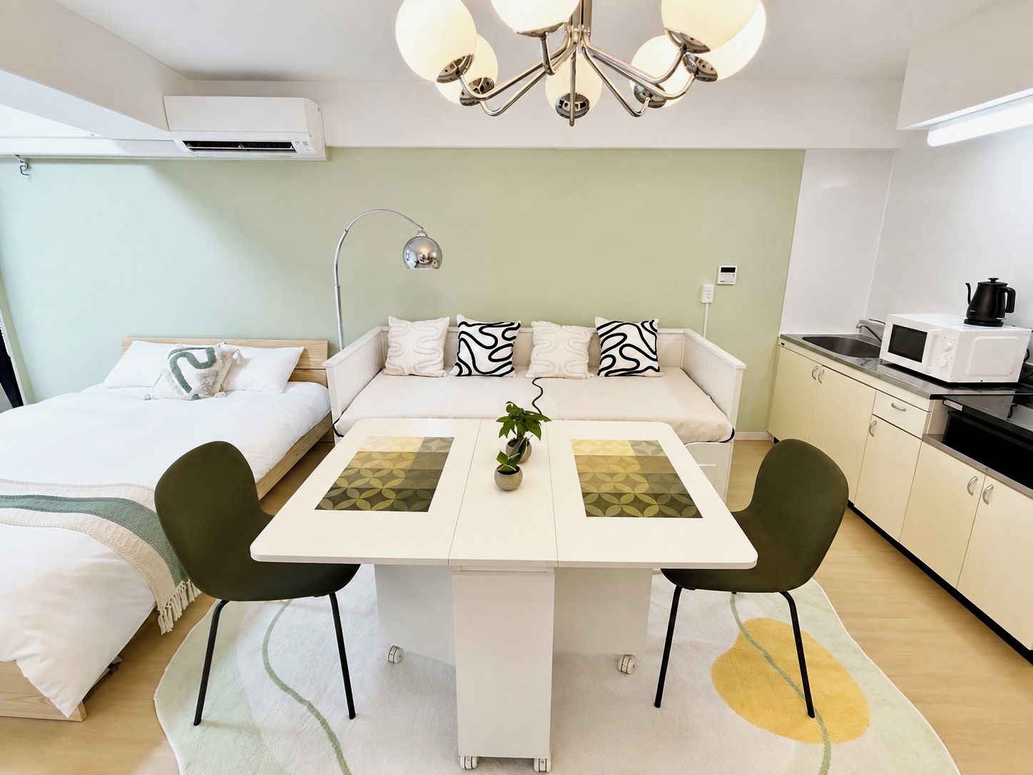 Een compacte kamer met een bed, een slaapbank, een eettafel met stoelen, een kitchenette en een plafondlamp.