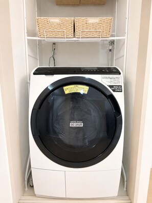 Eine Waschmaschine mit Frontlader und Bedienfeld, in einem Waschraum mit Regalen darüber.