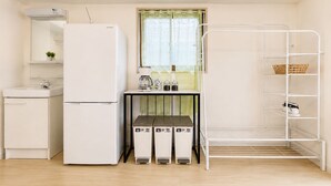 Cocina compacta con refrigerador, pileta y ventana con cortina. Se observa una estantería metálica con cestas y una pequeña mesa con hervidor y frascos.