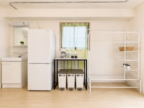 Eine kleine Küche mit Kühlschrank, Spüle und einem Fenster mit Vorhang. Es ist ein Metallregal mit Körben und ein kleiner Tisch mit Wasserkocher und Gläsern.