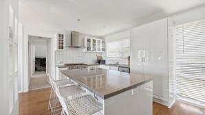 Een moderne keuken met witte kasten, een groot eiland met barkrukken, een fornuis en een raam met jaloezieën.