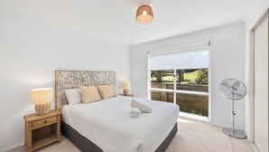 Een slaapkamer met een groot bed, twee nachtkastjes met lampen, een staande ventilator en een raam met uitzicht op groen.