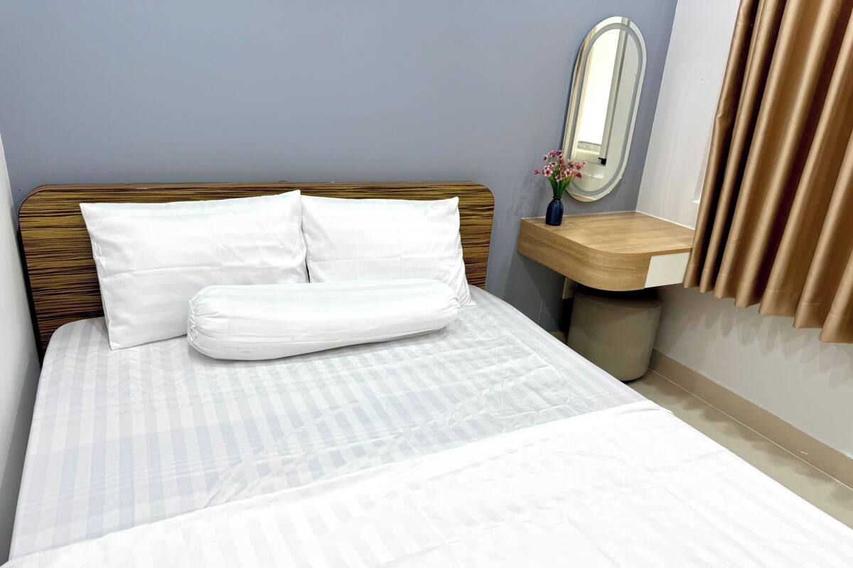Ein ordentlich bezogenes Bett mit weißen Leinen, ein Holz-Kopfschild, ein kleiner Nachttisch mit Blumen in einer Vase und ein Spiegel an der Wand.