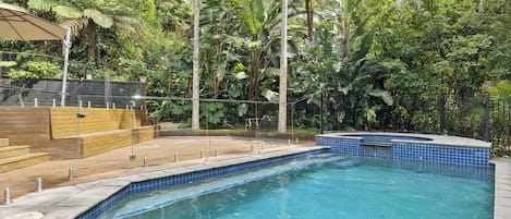 Une piscine dont le bord est revêtu de carrelage, entourée d’une végétation luxuriante et dotée d’une terrasse en bois.