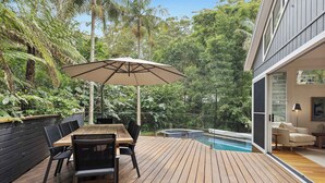A wooden deck with a table and chairs, an umbrella, and a pool in the background. - Pool & Spa Oasis. Home Theatre. Sleeps 8 (North Avoca)