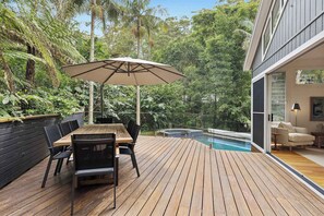 Eine Holzterrasse mit Tisch, Stühlen und einem Sonnenschirm, im Hintergrund ein Pool.