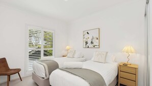A bedroom with two beds, a chair, a nightstand with a lamp, and a window with blinds. - Pool & Spa Oasis. Home Theatre. Sleeps 8 (North Avoca)