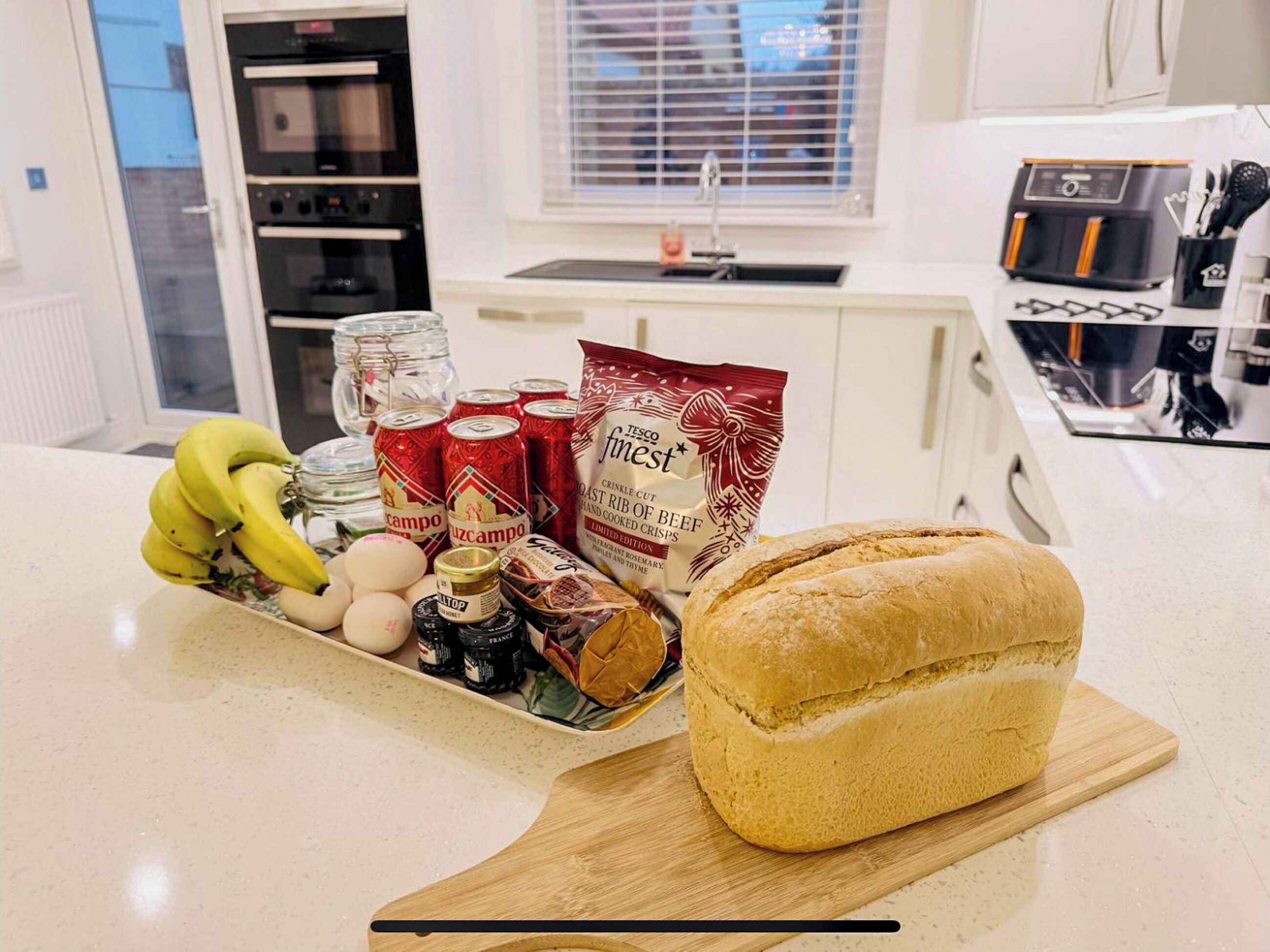 Una encimera de cocina con hogaza de pan, plátanos, huevos, bebidas en lata y una bolsa de papas fritas.