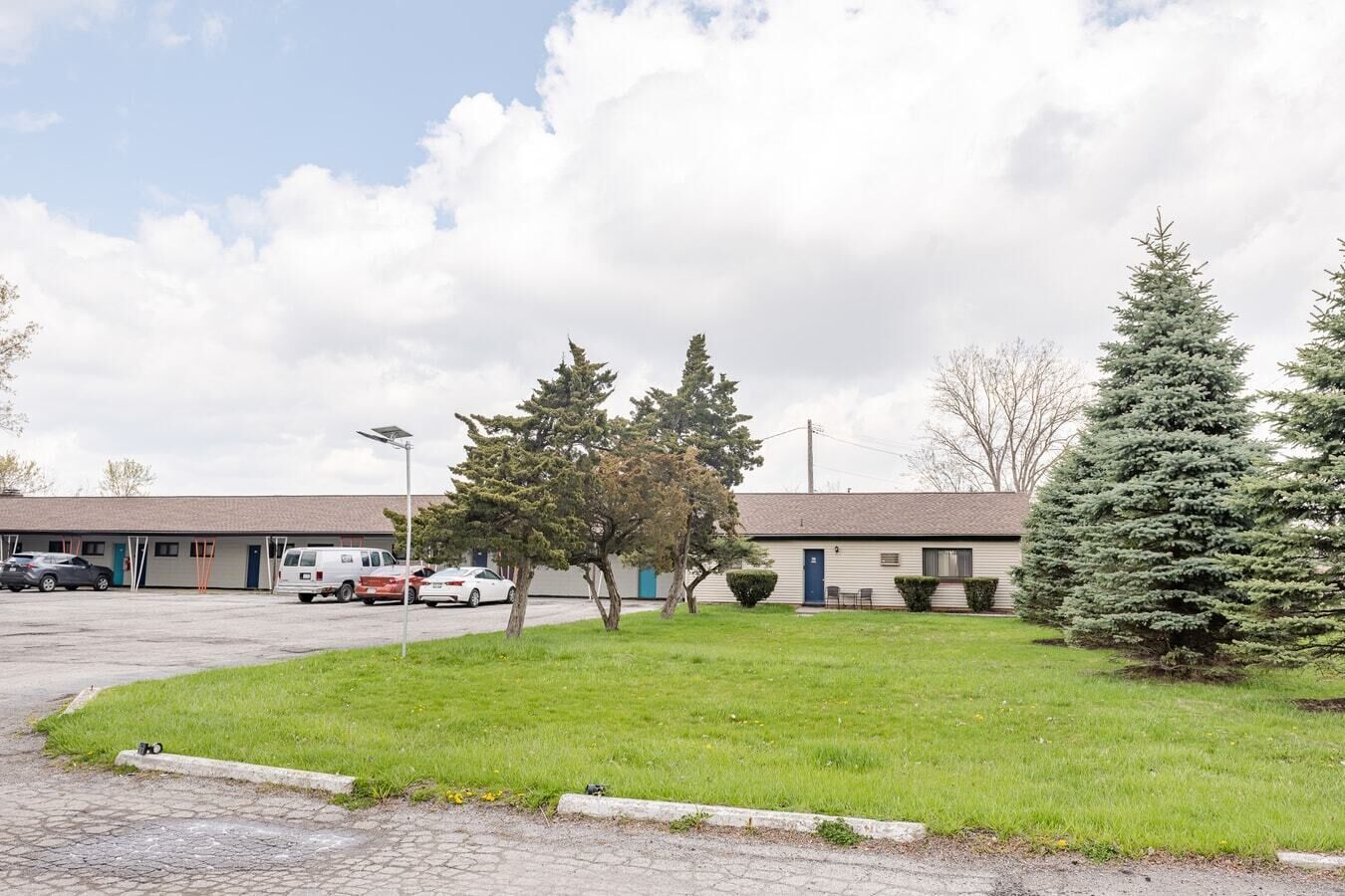Un motel avec un parking, plusieurs voitures et deux arbres à feuilles persistantes au premier plan.