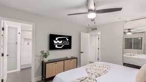A modern bedroom with a bed, a ceiling fan, a television displaying 'WAA PHOENIX OPEN', and a bathroom visible through an open door. - Old Town Dream- Pool, Hot Tub, Firepit, Foosball, Cornhole, and Mini Golf (Scottsdale)