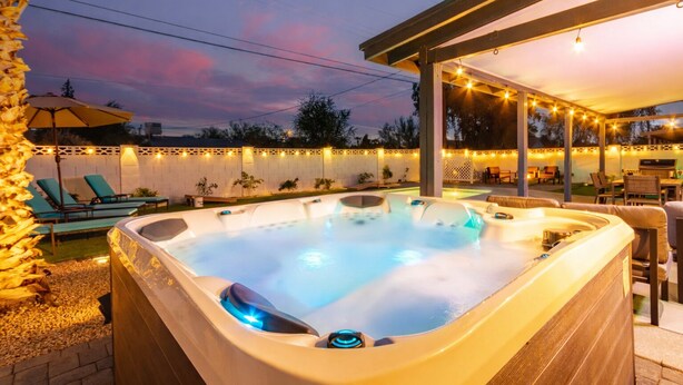 A hot tub with a wooden deck and string lights, surrounded by lounge chairs and a covered patio area. - Old Town Dream- Pool, Hot Tub, Firepit, Foosball, Cornhole, and Mini Golf (Scottsdale)