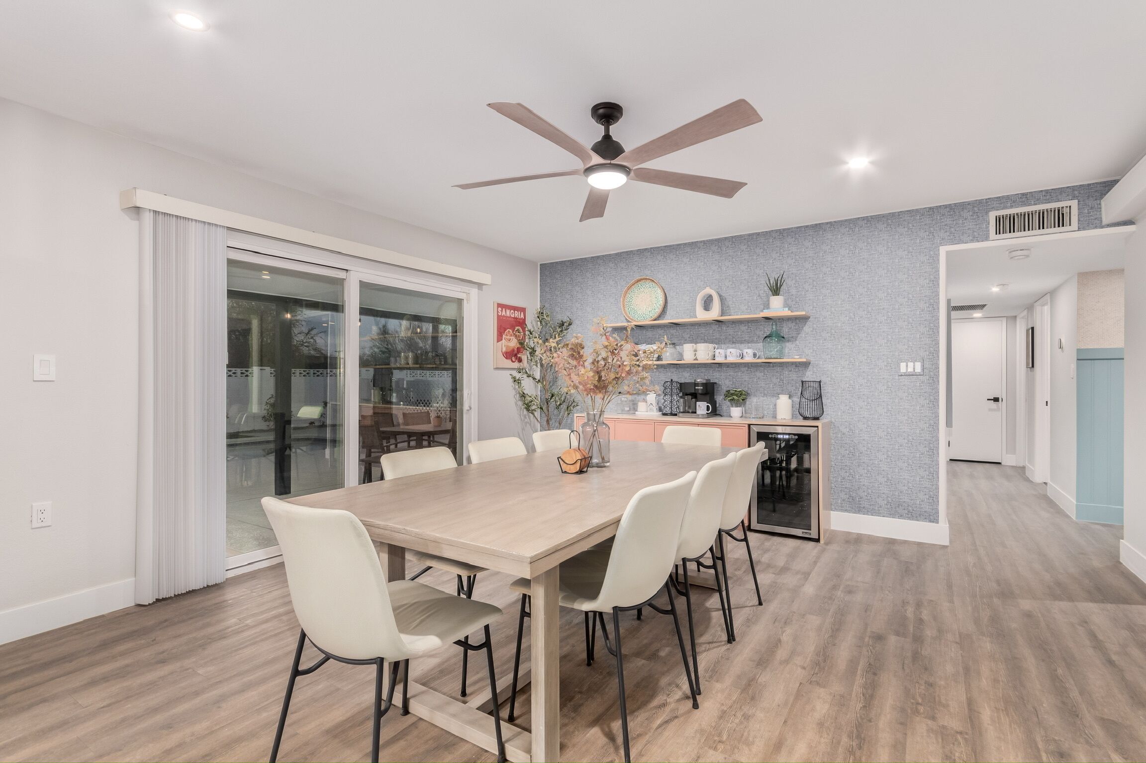 Un espace repas moderne avec une table en bois, des chaises blanches, un ventilateur de plafond et un coin cuisine doté d’étagères et d’objets décoratifs.