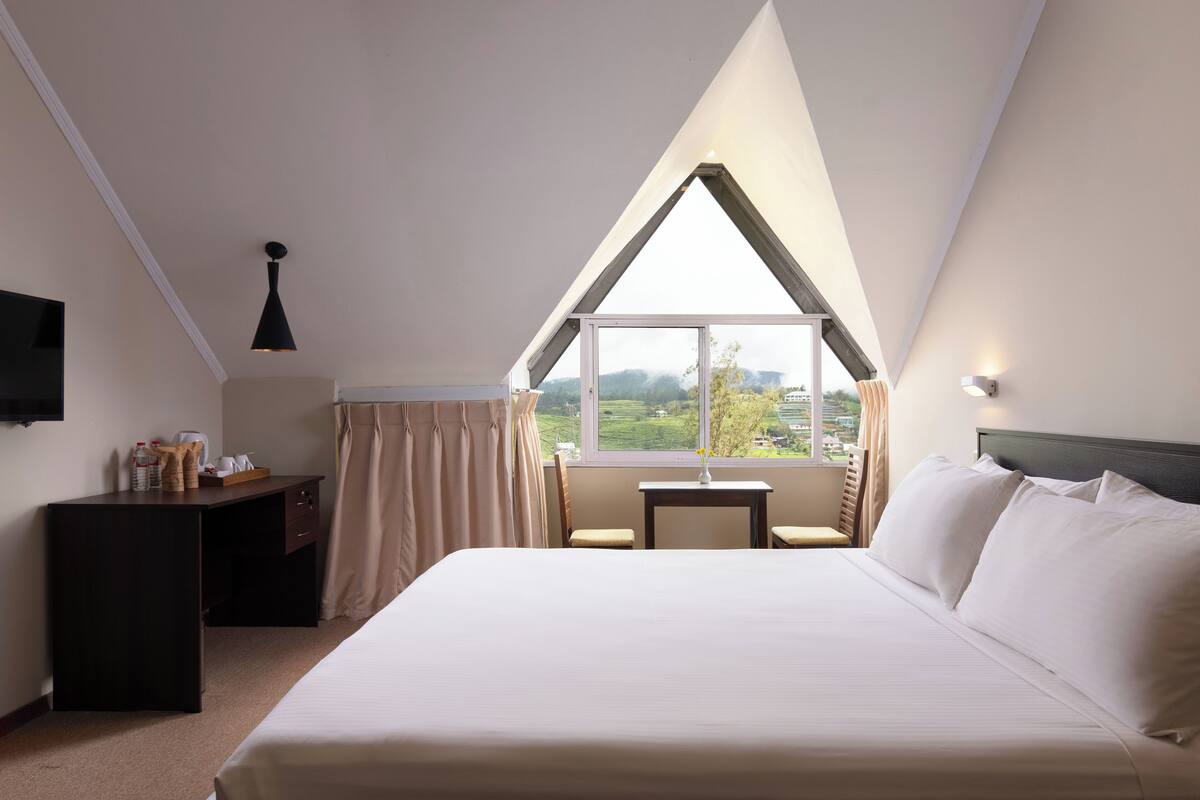 A hotel room with a large bed, a desk, a chair, a small table, and a window with a view of the countryside.