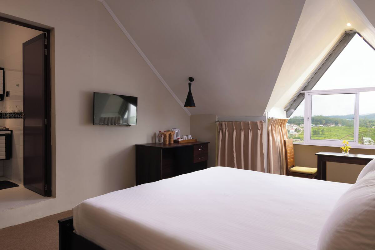 A bedroom with a large bed, a desk, a chair, a TV, and a window with a view of the countryside.