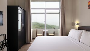 Habitación de hotel con una cama grande, un escritorio, dos sillas, un armario y una ventana con vistas a un paisaje verde.