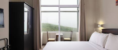 A hotel room with a large bed, a desk, two chairs, a wardrobe, and a window with a view of a green landscape.