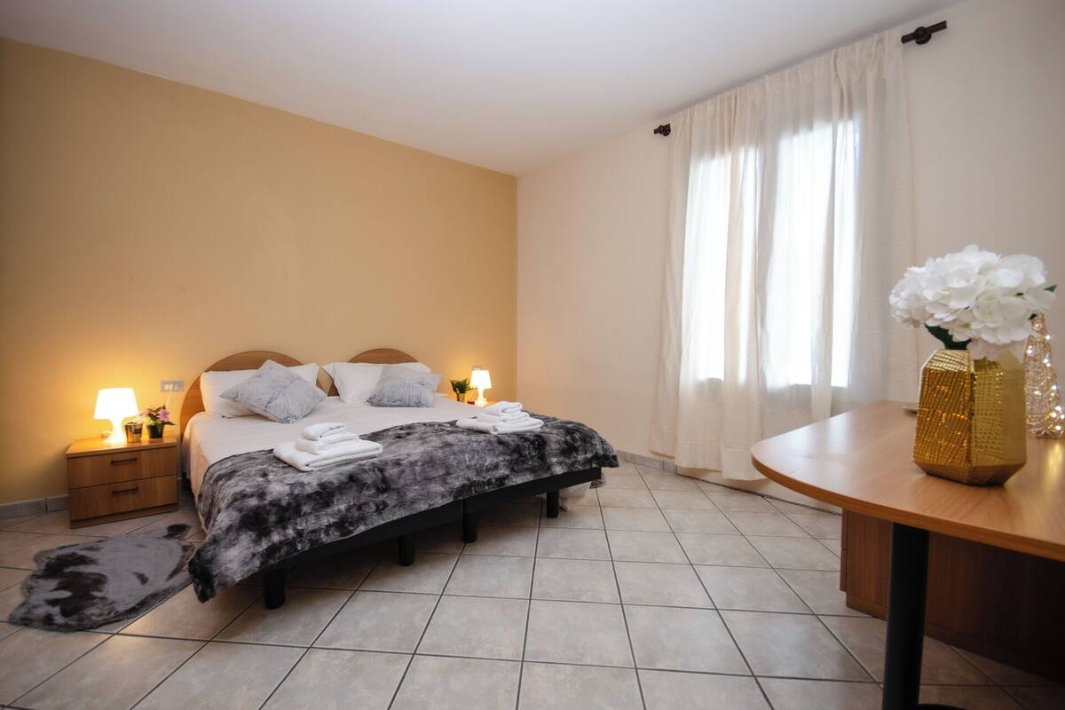 A neatly arranged bedroom with a bed, bedside tables, a lamp, a rug, a table with a vase, and a window with curtains.