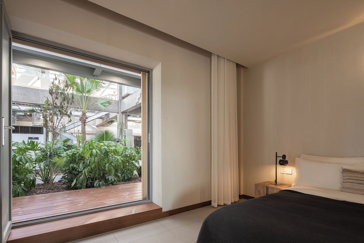 Habitación de hotel moderna con un amplio ventanal corredero que da a un balcón exterior con plantas y vistas del interior del edificio.