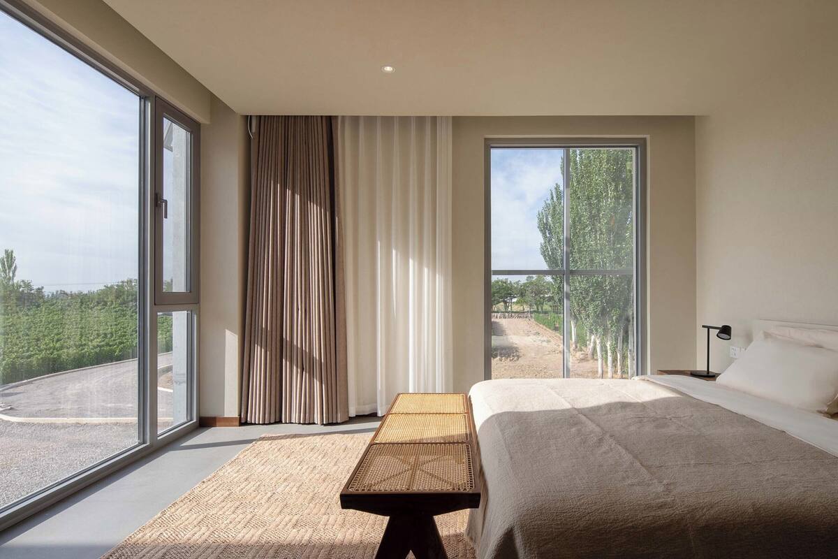 Un dormitorio con una cama grande, una mesita de noche de madera y vistas a una carretera y árboles a través de amplias ventanas.