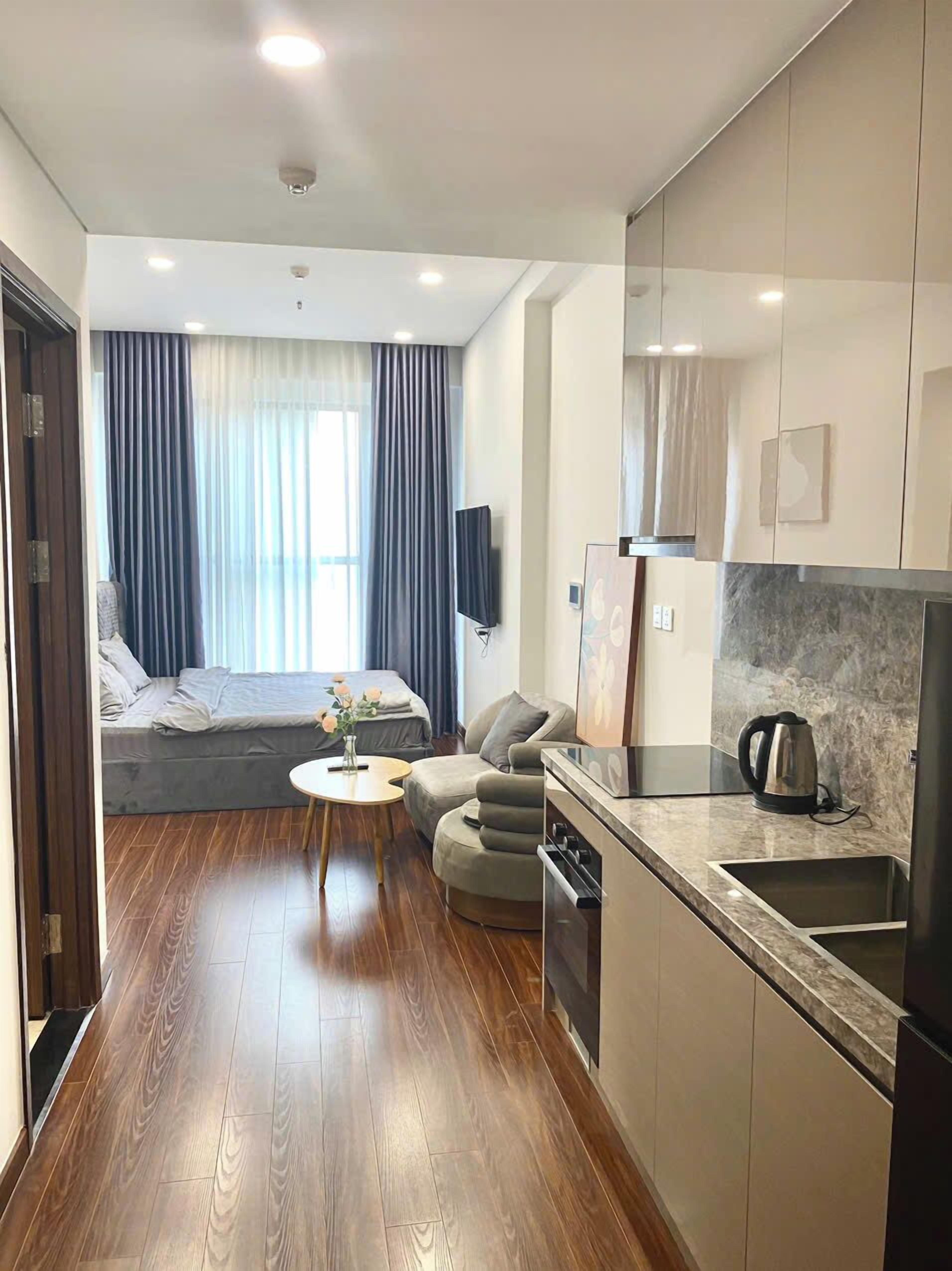 Una habitación de hotel moderna con suelo de madera, un sofá cama, una mesita redonda pequeña, una kitchenette con un fregadero y hervidor, y un televisor montado en la pared.