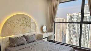 A modern bedroom with a large bed, a bedside table with a mirror, and a view of high-rise buildings. - Vinhomes Ocean Park 1 Gia Lâm Apartment (Hanoi)