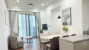 A modern living room with a sofa, dining table, and chairs. There is a large window with curtains, a flat-screen TV, and a plant on the dining table. - Vinhomes Ocean Park 1 Gia Lâm Apartment (Hanoi)