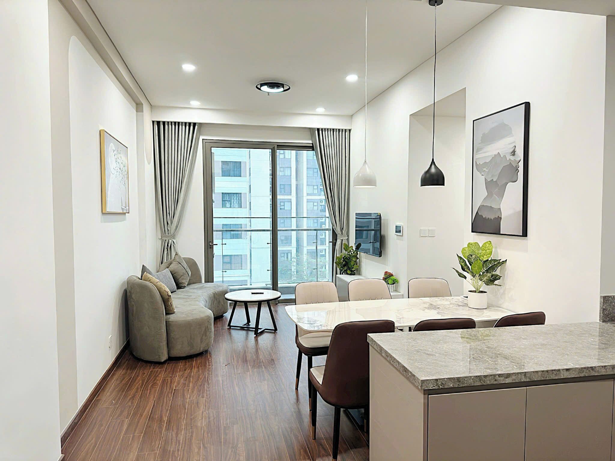Un salon moderne avec un canapé, une table à manger et des chaises. On y trouve une grande fenêtre avec des rideaux, un luminaire au plafond et un tableau accroché au mur.