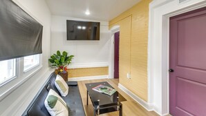 A room with a black sofa, a small black table with a magazine, a TV on the wall, a window with a blind, and a door with a purple frame. - Colorful & Convenient Darby Apt, 7 Mi to Stadiums! (Darby)