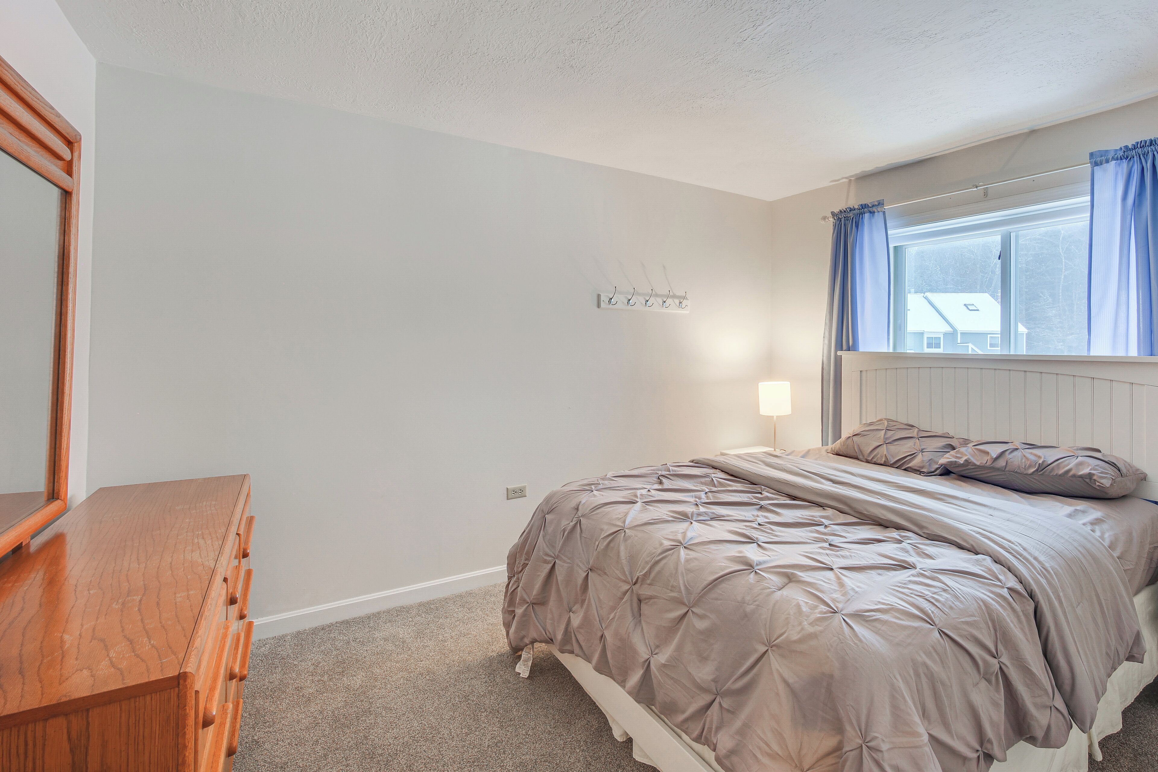 A bedroom with a wooden dresser, a bed with a beige quilt, a window with blue curtains, and a wall-mounted light fixture.