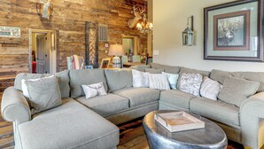 A spacious living room with a large sectional sofa, a round coffee table, and a wooden accent wall. - 4,800-Sq-Ft Cabin w/ Creek Access in Caulfield (Caulfield)