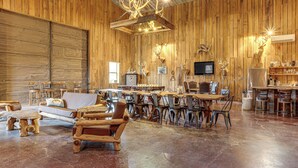 A spacious room with wooden walls and floors, featuring a large dining table, several chairs, a sofa, and a fireplace. - 4,800-Sq-Ft Cabin w/ Creek Access in Caulfield (Caulfield)
